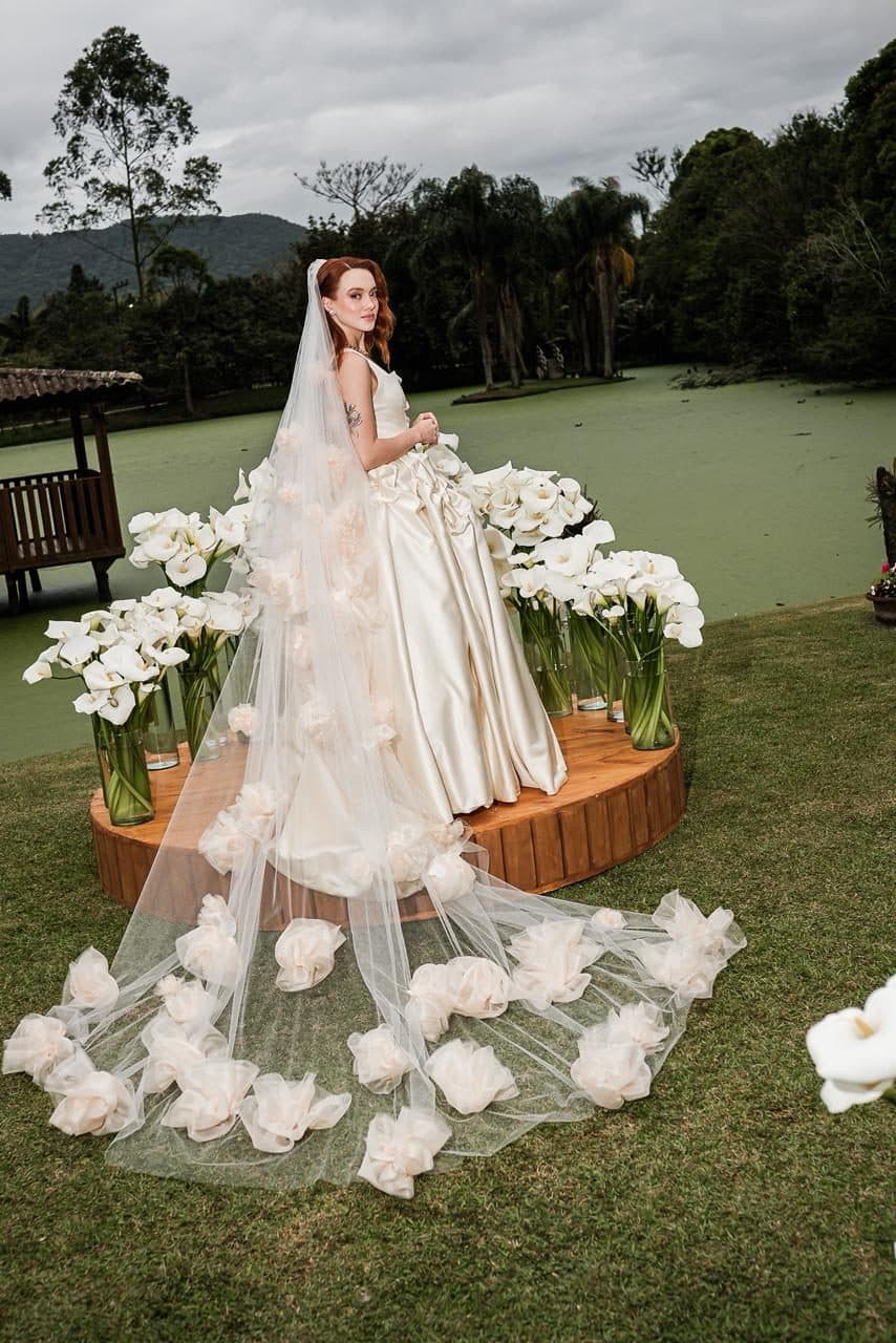 Noiva ruiva em um jardim, com véu longo coberto de flores, ao lado de um lago